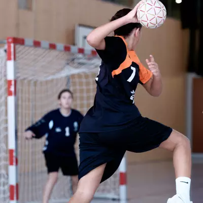 A player in a black and orange kit preparing to take a shot in handball, facing the goalkeeper in front of the goal.