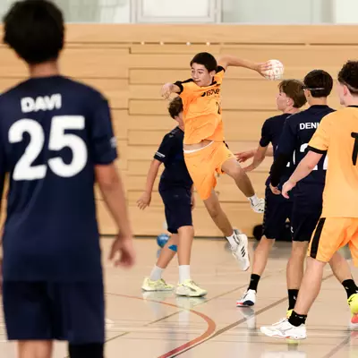 Handball player in an orange jersey jumping to take a shot, surrounded by teammates and opponents in navy blue on the court.
