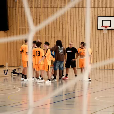 Players in orange jerseys gathered around their coach on a handball court, seen through the netting of a goal.