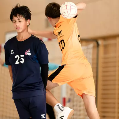Two handball players face off, one in an orange jersey jumping to shoot, the other in navy blue number 25 watching.