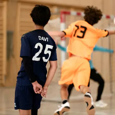 A player in orange, number 13, jumps to take a shot at goal, while a defender in navy blue, number 25, looks on.