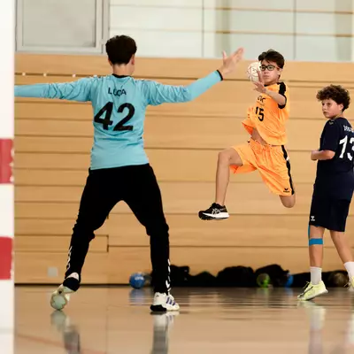 A goalkeeper in cyan tries to save a shot from a player in orange jumping in the air, while another player in navy blue looks on.