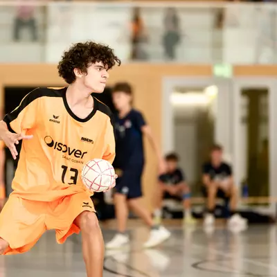 Young player in an orange number 13 shirt holding a handball on the court, with opponents in the background.
