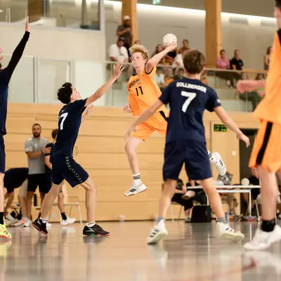 Handball player in orange jumping to take a shot, surrounded by defenders in navy blue on an indoor court.