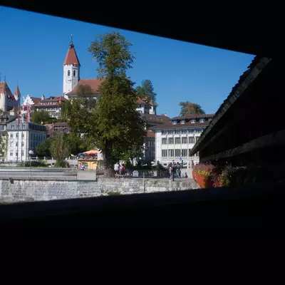 View of Thun's old town from a covered wooden bridge, with a castle tower, church spire, and pastel buildings under a clear sky.