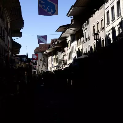 Narrow street in Thun with tall silhouetted buildings and a purple bear-crest flag hanging overhead against a clear blue sky.