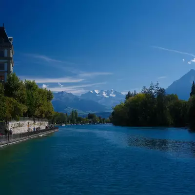 A calm river flows through Thun, lined with trees and a historic building, with snow-capped mountains in the distance.