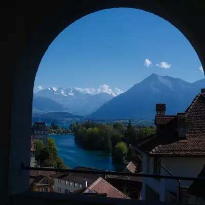 View through a stone archway of Thun's river, red-tiled rooftops, and mountains under a blue sky.