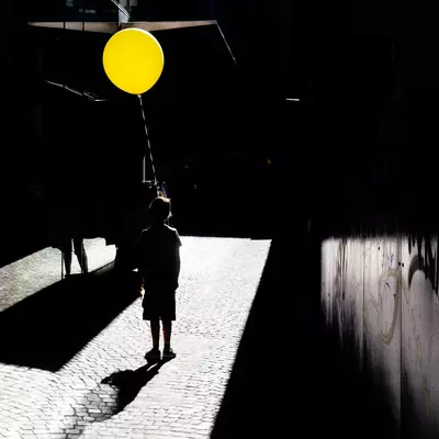 Silhouette of a child holding a yellow balloon, standing on cobblestones lit by a beam of light in a dark alleyway.