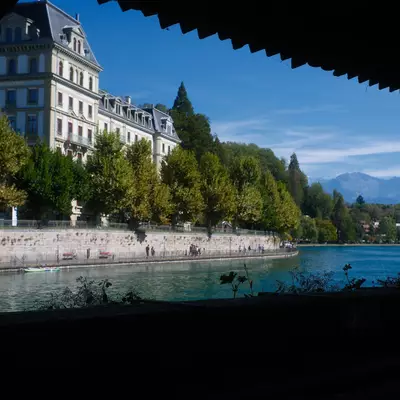 View from beneath a wooden roof overhang of the Aare river in Thun, lined with trees and a grand historic building, mountains in the distance.