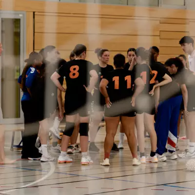 Joueuses de handball en maillots noirs (numéros 8, 11, 12) réunies en cercle sur le terrain, avec un coach à proximité.