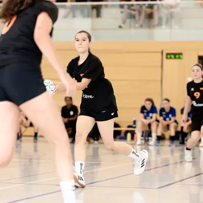 Joueuses de handball en action sur le terrain, l'une en noir portant le ballon, poursuivie par une joueuse en bleu au second plan.