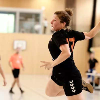 A female handball player in a black jersey jumps mid-air to throw the ball, with a referee and opposing players nearby.
