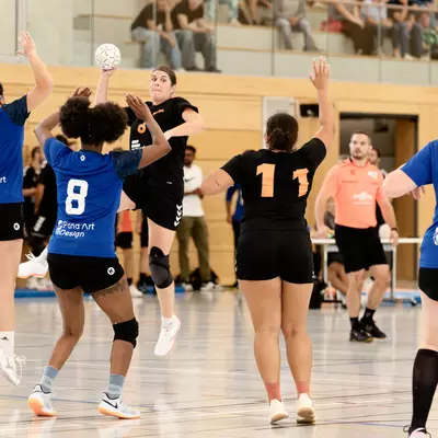 Joueuse en noir sautant pour tirer, entourée de joueuses en bleu bras levés, lors d'un match de handball en salle.