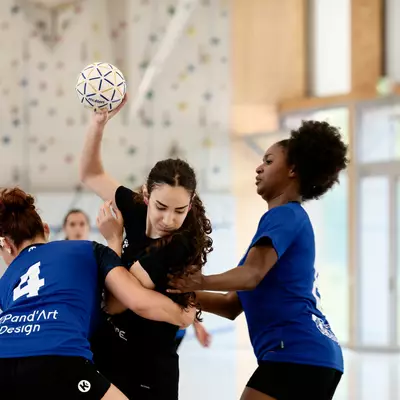 A player in black holds the ball high above two opposing players in blue during an indoor handball match.