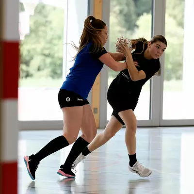Handball player in black holding the ball while being challenged by a defender in blue on an indoor court.