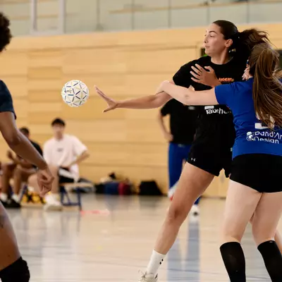 Joueuse en noir passant le ballon face à deux joueuses en bleu lors d'un match de handball en salle.