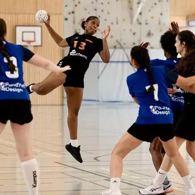 Joueuse de handball en noir (n°13) sautant pour tirer, entourée de joueuses en bleu sur un terrain intérieur.