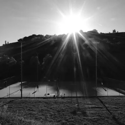 Black-and-white photo of tennis courts backlit by a bright sun, with tall trees and hills silhouetted in the background.