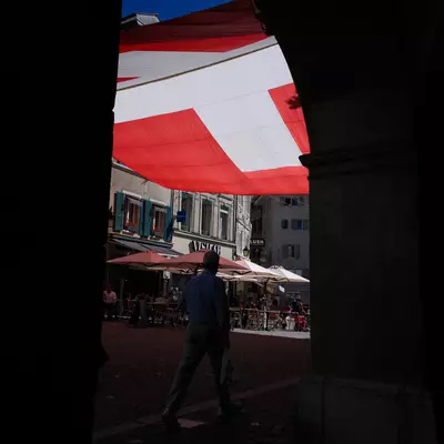 A large Swiss flag stretches across a stone archway, framing a pedestrian walking through the sunlit square below.