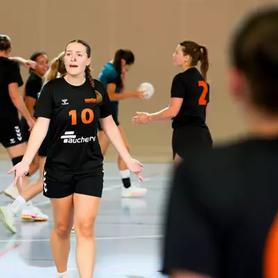 Handball players in black jerseys with orange numbers stand on an indoor court.