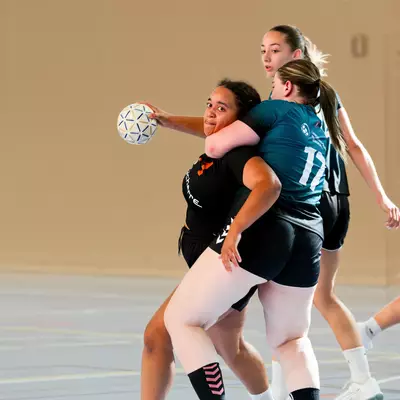 Handball players in black and teal jerseys contest possession of the ball on an indoor court during a match.
