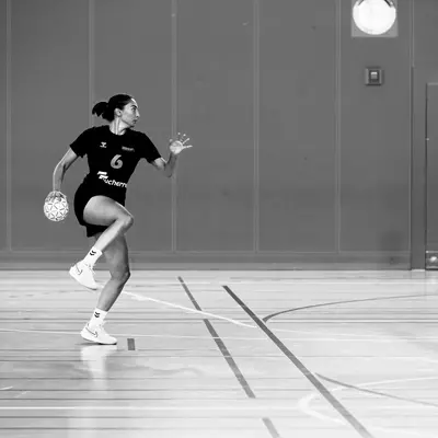 Handball player wearing number 6 dribbles the ball across the court, with a teammate and referee watching in the background.