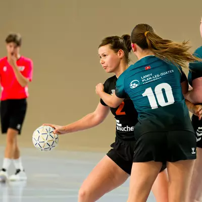 Handball players in teal jerseys numbered 10 and 17 defend against an opponent dribbling the ball on court.