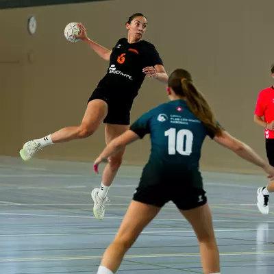 A handball player in a black jersey leaps mid-air to shoot, while a defender in a navy jersey number 10 blocks.