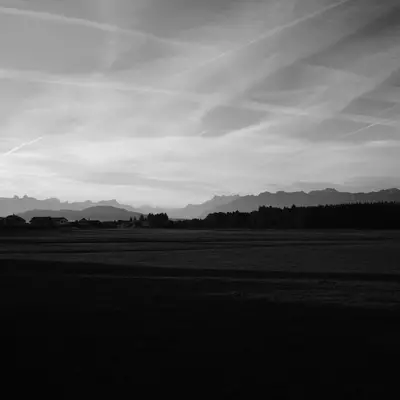 Photo of a panorama in black and white. In the distance, we can see the Alps standing out against a sky streaked with straight-lined clouds.