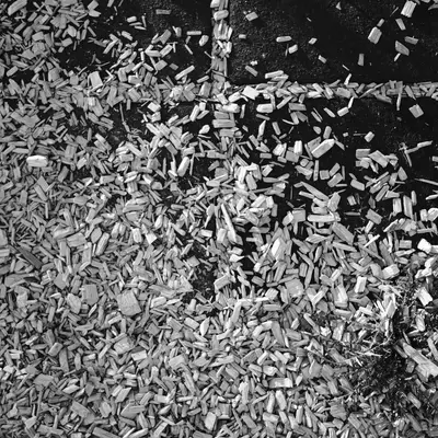 Black-and-white photograph of the ground. It is a playground surface made of impact-absorbing tiles, which are largely covered with wood chips.