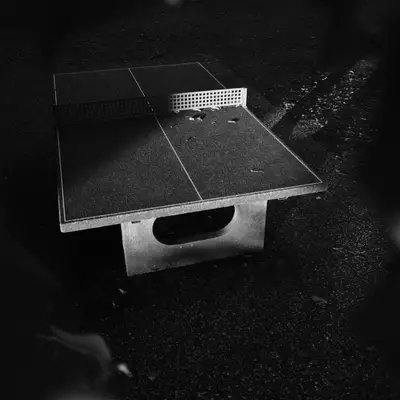 Black and white photo. It is a ping-pong table lit diagonally by the sun. Only the bottom right quarter, as well as half of the net, are lit, the rest is in the shadows.