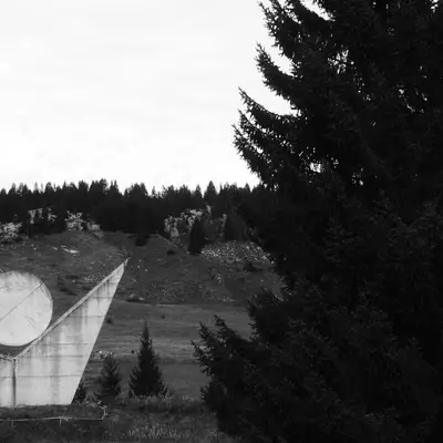 Black and white photo of a triangular concrete monument topped with a circle, in a hilly landscape lined with fir trees.
