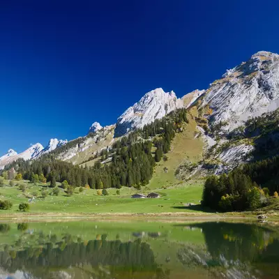Jagged mountain peaks and green forested slopes near La Clusaz reflected in a calm alpine lake under a clear blue sky.