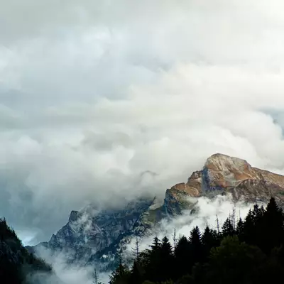 Rocky mountain rising out of thick clouds, surrounded by conifer forests, in the La Clusaz area.