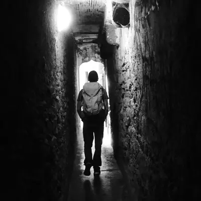 Silhouette of a person with a backpack walking through a narrow stone passage toward a bright exit.