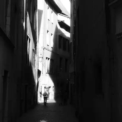 Narrow black and white alley lined with tall buildings, with a figure walking in the distance under a ray of light.