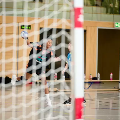 Handball player in mid-air throwing the ball toward the goal, viewed through the net, during an indoor match.