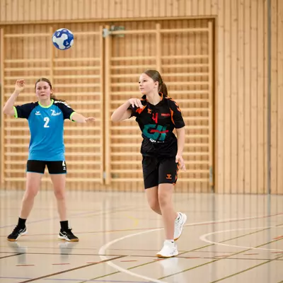 Young handball players in a gymnasium, one in black jersey tracking a thrown ball, teammates in blue nearby.