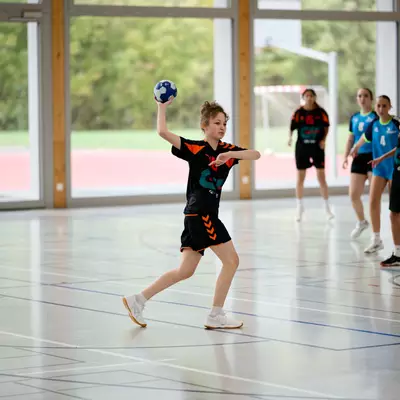 Young handball player in black and orange jersey throwing the ball, with opposing team members standing behind in defense.