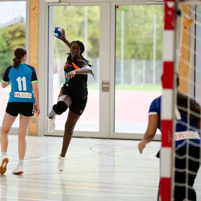 Female handball player leaping mid-air to shoot at the goal, with a defender and goalkeeper nearby on an indoor court.