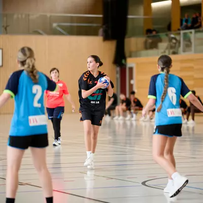 Young female handball players in blue and orange jerseys on an indoor court, with one holding the ball mid-play.