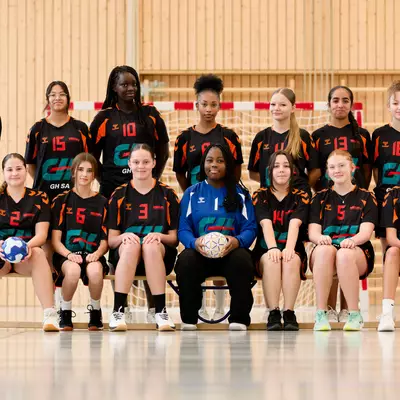 Team photo of young female handball players in orange and black jerseys, standing and seated in rows, with a coach and three holding handballs.