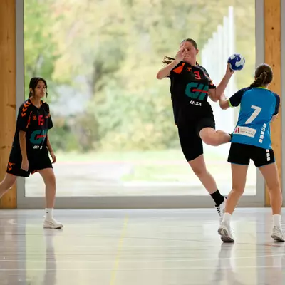 Handball player in black jumping to shoot while a defender in blue blocks, with a teammate watching nearby on an indoor court.