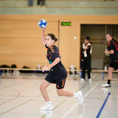 Young handball player in dark uniform running with raised arm holding the ball, preparing to throw during a match.