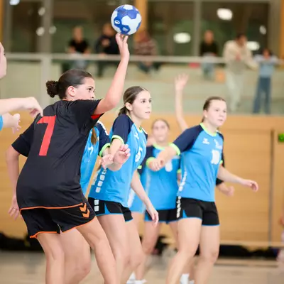 Handball player in black jersey number 7 jumps to shoot while defenders in light blue surround her on the court.