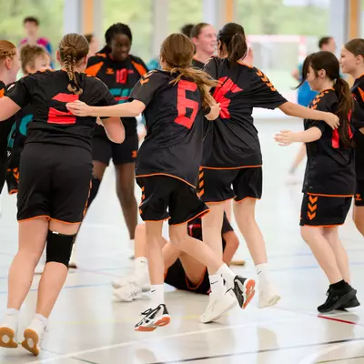 A youth handball team in black and orange jerseys celebrates together in a huddle on an indoor court.