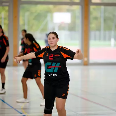 Young handball players in black and orange jerseys practice on an indoor court, with one girl extending her arms in focus.