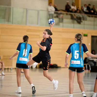 Young female handball player leaping to throw the ball, with teammates and opponents in blue and black jerseys on the court.