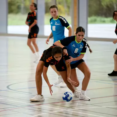 Young female handball players in black and blue jerseys compete for the ball on an indoor court during a match.